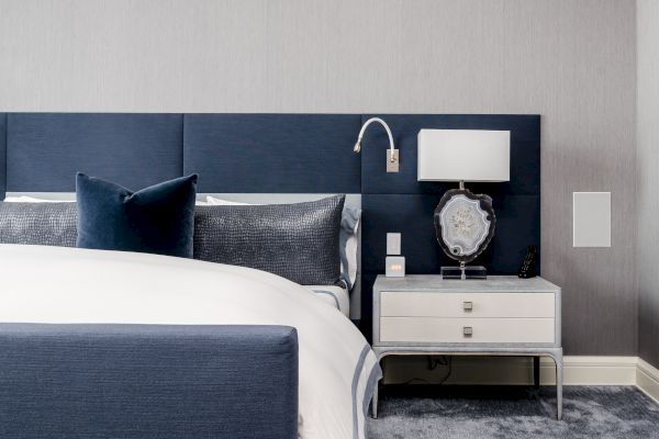A modern bedroom with a blue bed, grey and white pillows, a white nightstand, a lamp, and a decorative object on the nightstand.
