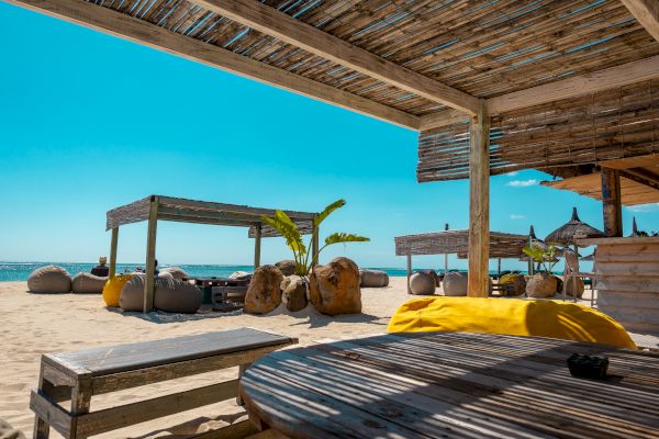 A sunny beach scene with shaded seating areas, wooden structures, and cozy bean bags on sandy shores; clear blue sky overhead.