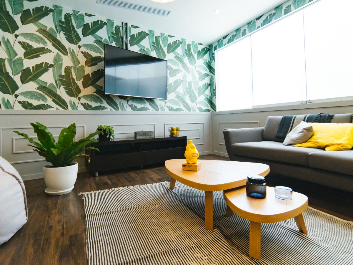 A modern living room with tropical wallpaper, a gray sofa, wooden coffee tables, a TV, a rug, and indoor plants, featuring yellow accents.