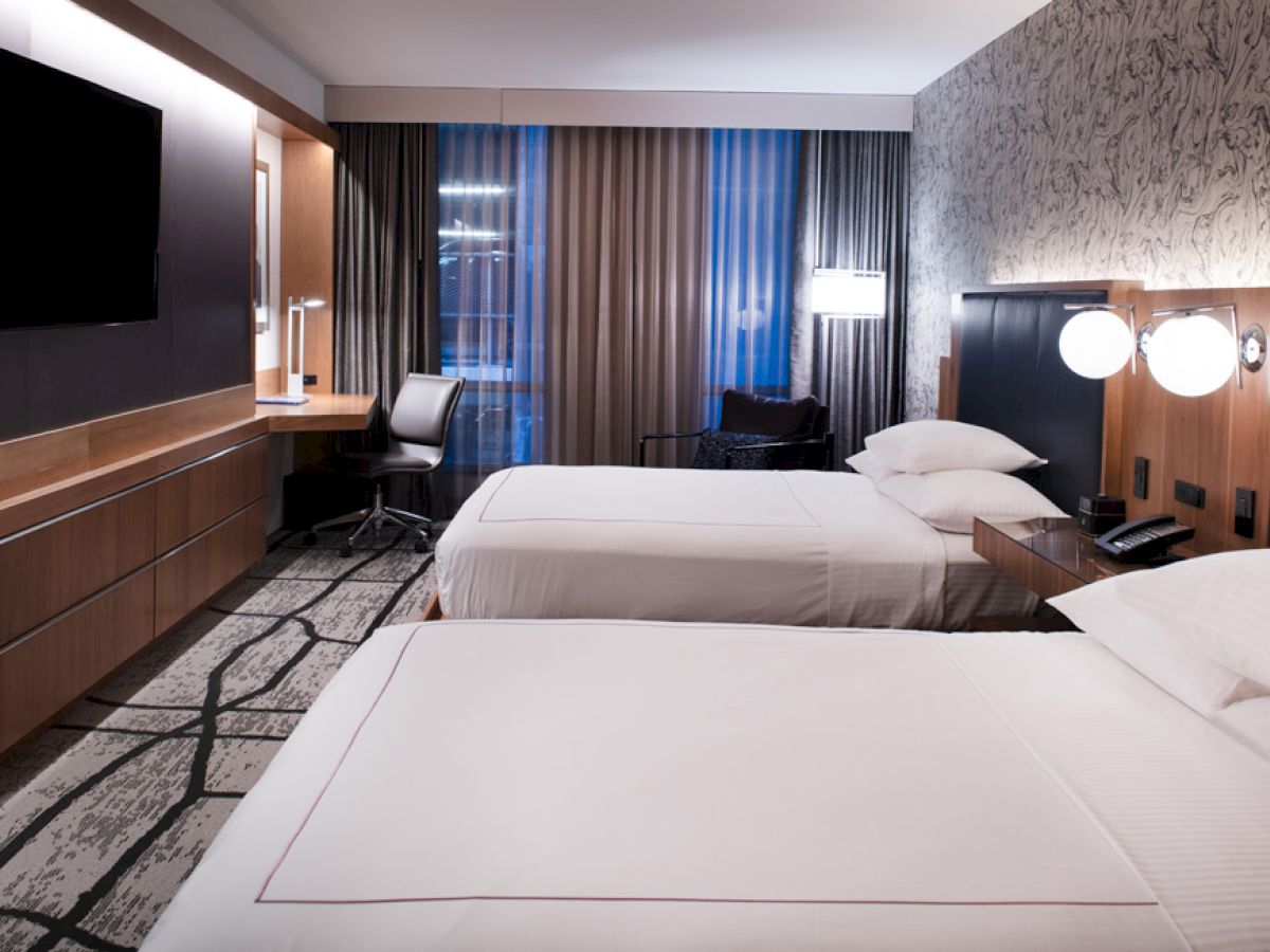 A modern hotel room with two double beds, a large TV, a desk with a chair, and contemporary decor, featuring stylish wallpaper and lamps.