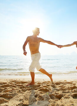 A man and woman hold hands while running on a beach by the ocean, basking in the sunlight on a clear day.