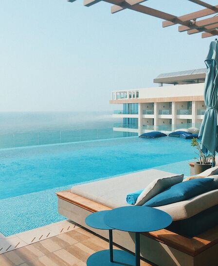 An infinity pool with lounge chairs, umbrellas, and a sea view, framed by modern architecture on a sunny day.