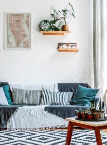 A cozy living room with a sofa adorned with pillows, plants, a coffee table, large windows, and wall art, creating a bright and inviting ambiance.