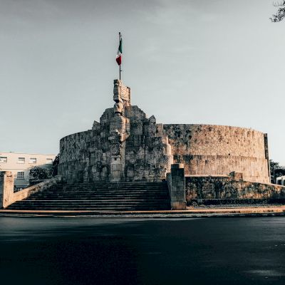 The image shows a monument with stone carvings and a circular base, topped by a flag. It is situated at the end of a set of stairs.