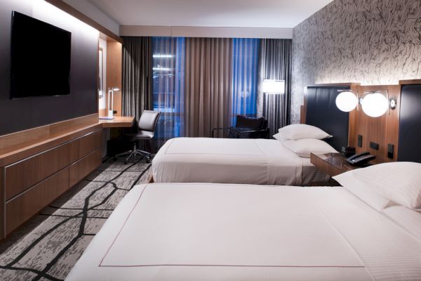 The image shows a modern hotel room with two neatly made beds, a TV, a work desk, and floor-to-ceiling windows covered with curtains.