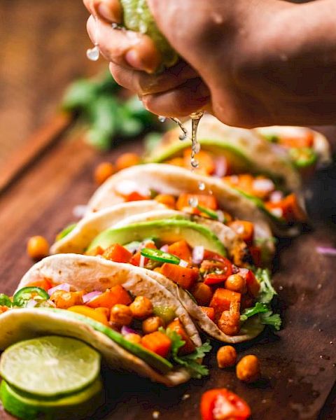 Five tacos are lined up on a wooden surface, filled with chickpeas, vegetables, and garnished with cilantro while lime juice is being squeezed on them.