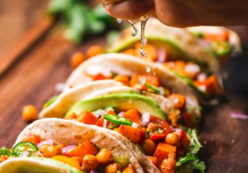 Five tacos are lined up on a wooden surface, filled with chickpeas, vegetables, and garnished with cilantro while lime juice is being squeezed on them.
