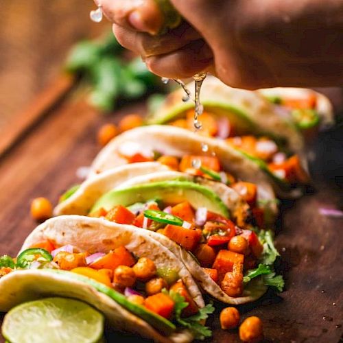 Five tacos are lined up on a wooden surface, filled with chickpeas, vegetables, and garnished with cilantro while lime juice is being squeezed on them.