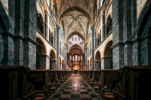 The image shows the interior of a grand cathedral with high vaulted ceilings, stone arches, and rows of wooden pews leading to an illuminated altar.