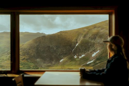 A person is sitting at a table, looking out a window at a vast, mountainous landscape with patches of snow.