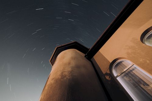 The image shows a night sky with star trails above a tall building with arched windows, captured from a low angle looking upwards towards the stars.