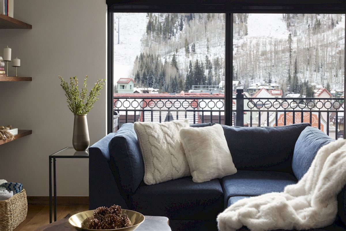 A cozy living room with a blue sectional sofa, white throws, shelves, fireplace, and large windows overlooking a snowy mountain village.