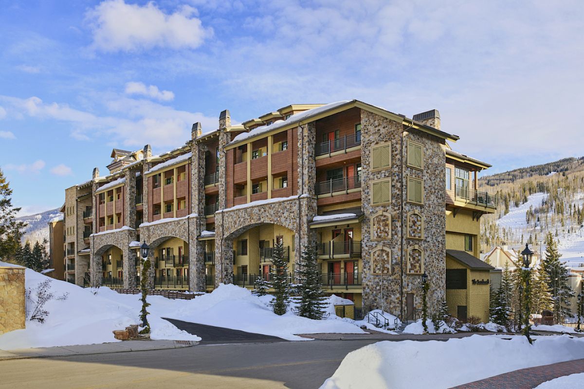 A large stone and wood building stands in a snowy mountain area with a clear blue sky and scattered clouds in the background.