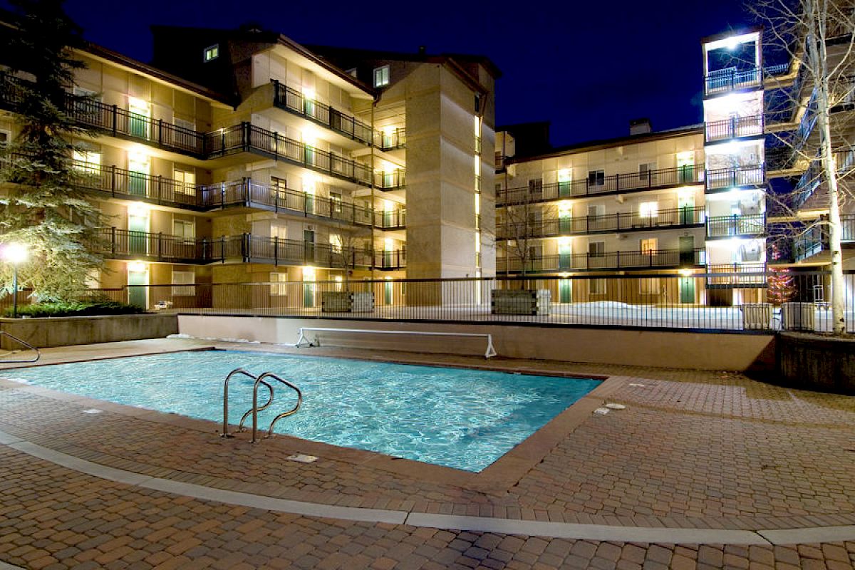 The image shows a lit outdoor swimming pool in front of a multi-story apartment building at night, surrounded by trees and outdoor lighting.