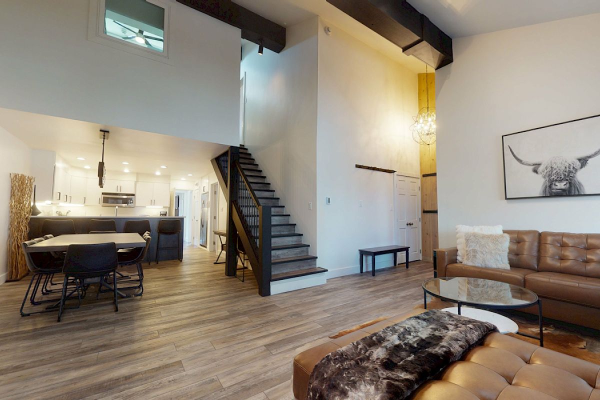A modern living space with an open floor plan, featuring a kitchen, dining area, and staircase. Decor includes a couch, artwork, and a chandelier.