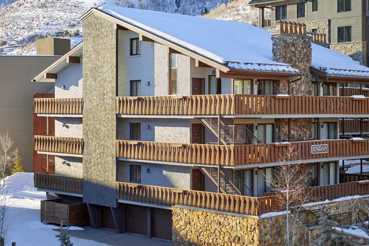 A snow-covered, multi-story building with wooden balconies. Other buildings surround it, set against a backdrop of snowy mountains and trees.