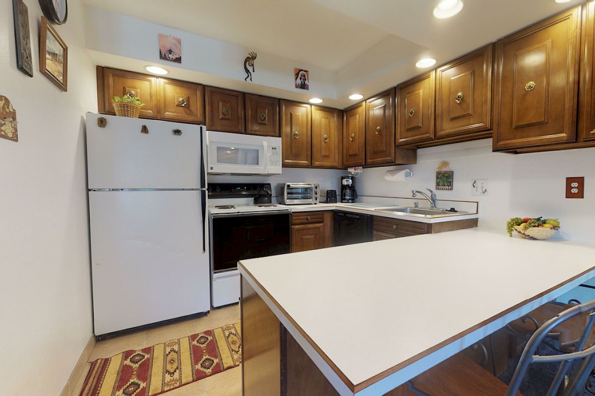 A cozy kitchen with wooden cabinets, white appliances, a countertop island, and a colorful rug on the floor. The lights are warm and inviting.