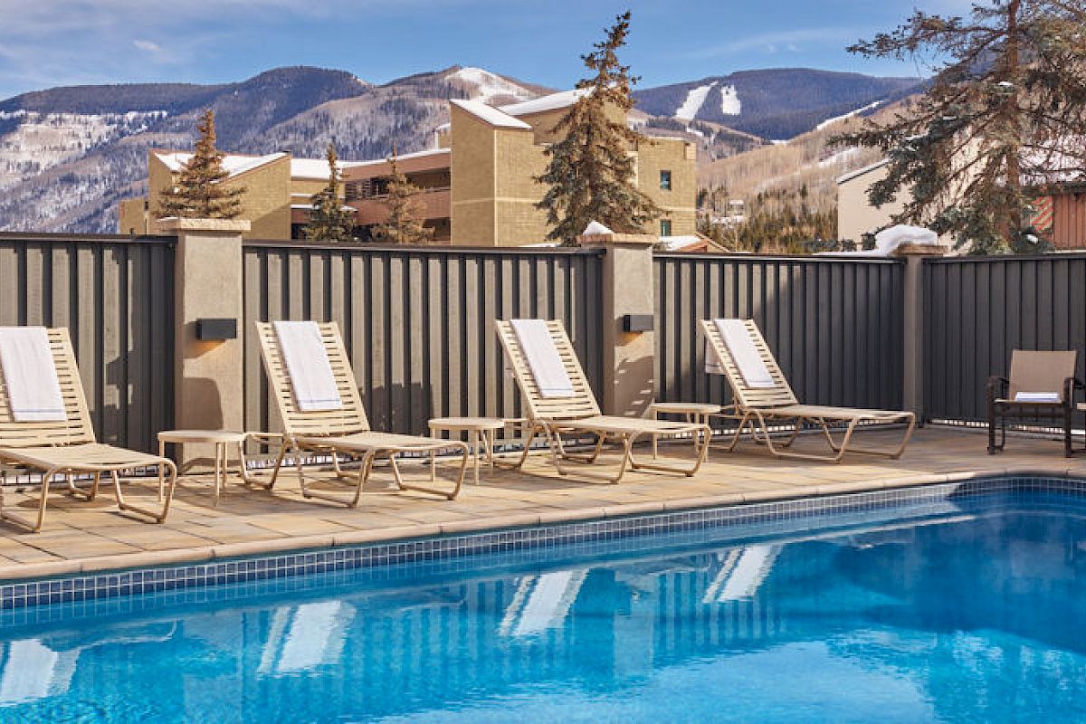 The image features an outdoor swimming pool surrounded by lounge chairs with mountains in the background under a partly cloudy sky.