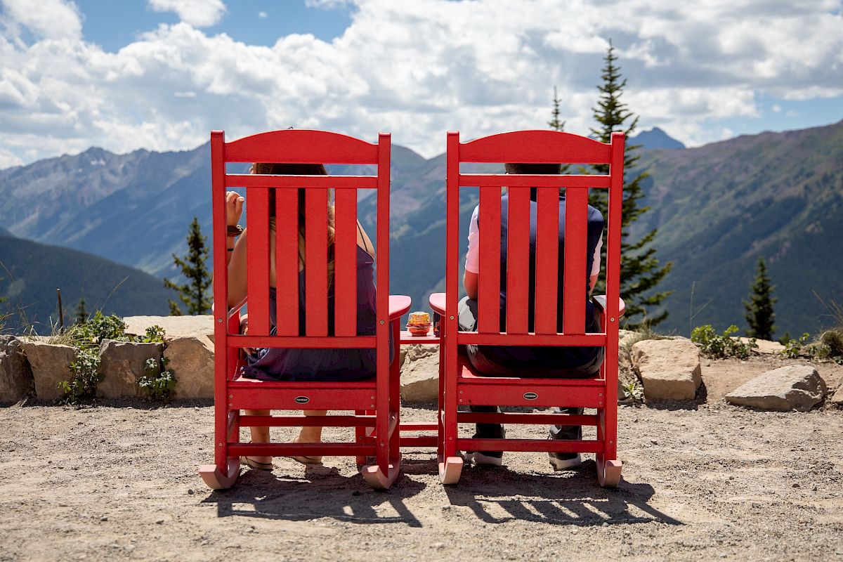 Two people are sitting in red rocking chairs, overlooking mountains and trees under a partly cloudy sky.