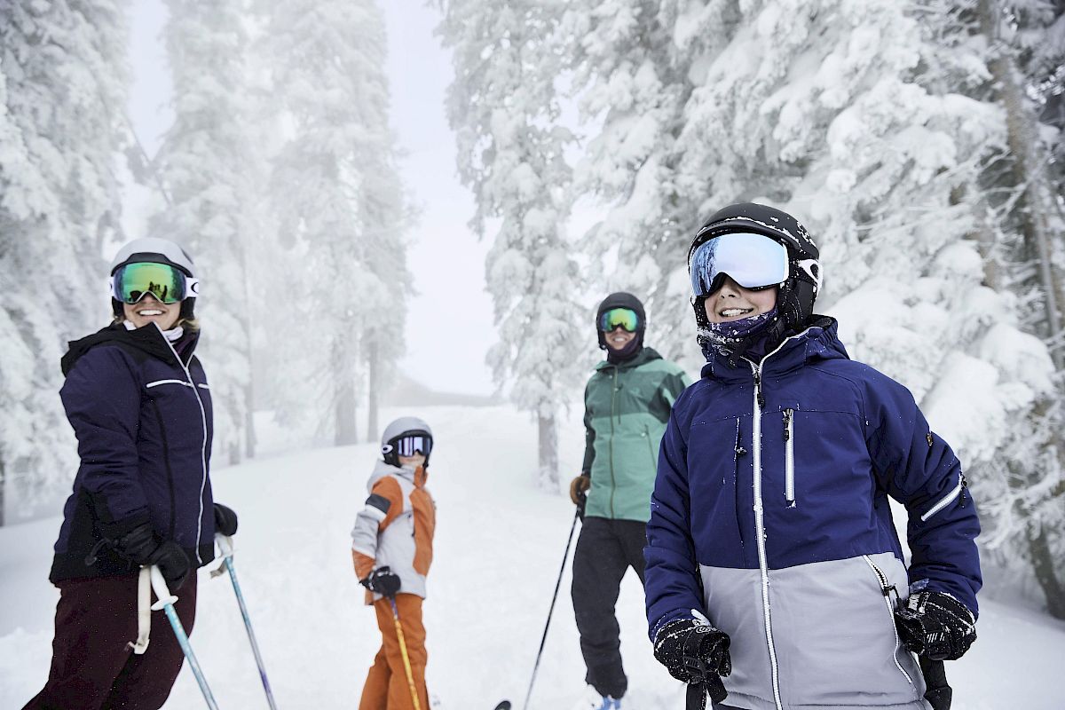 A group of four people in winter clothing and ski gear standing on a snowy slope surrounded by snow-covered trees.