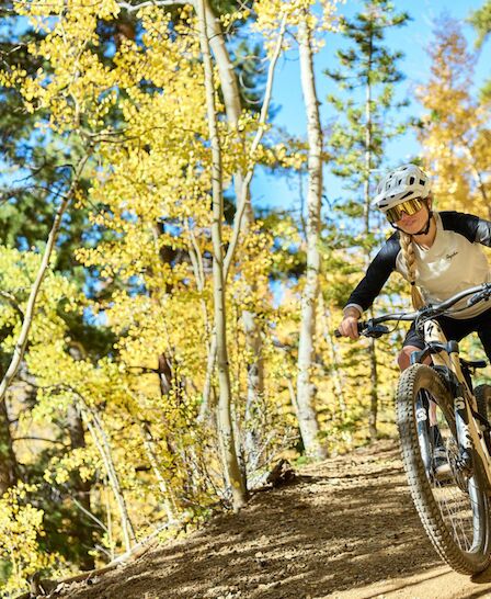 Two mountain bikers ride along a dirt trail surrounded by vibrant fall foliage and sunlight filtering through the trees.