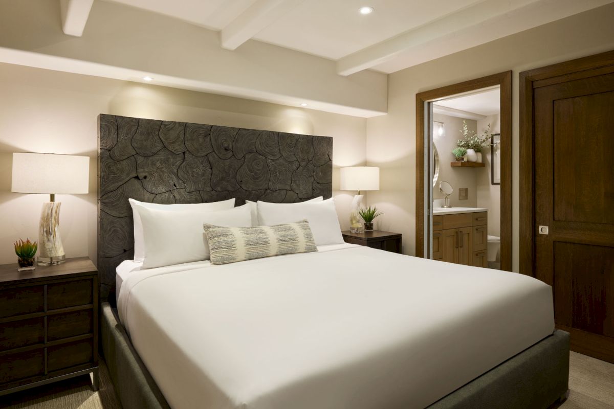 A cozy bedroom with a large bed, bedside tables with lamps, a cushioned headboard, and a view of an adjacent bathroom through an open door.