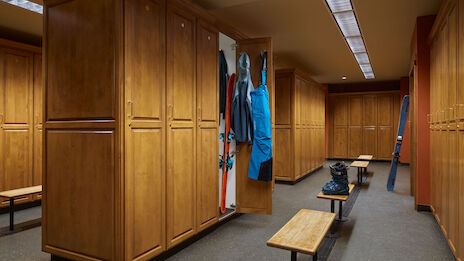A spacious locker room with wooden lockers, benches, and ski gear hanging. The floor is clean, and the ceiling lights are bright.