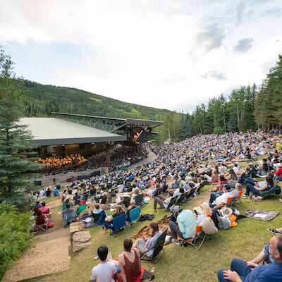 A large outdoor concert with a full audience sitting on a hillside, surrounded by trees and greenery under a partly cloudy sky.