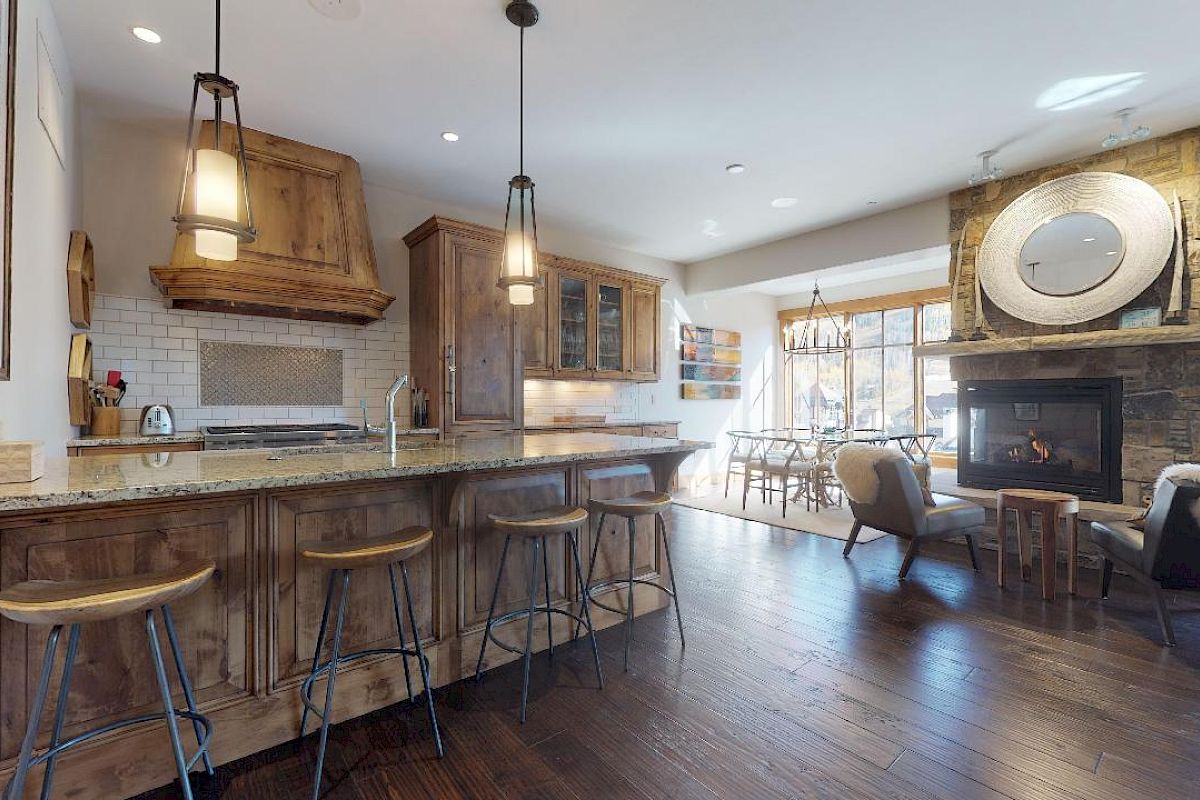 A cozy kitchen and living area with wooden cabinetry, island seating, a fireplace, and ample natural light through large windows with a dining space nearby.