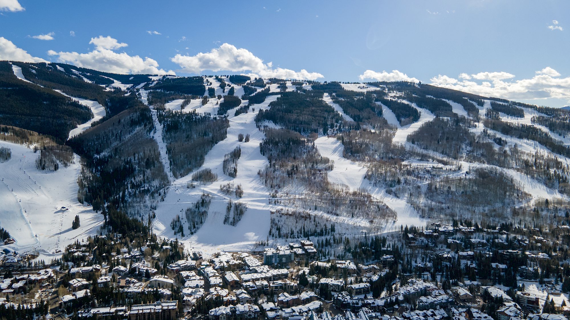 An aerial view of a snow-covered ski resort with numerous trails and a town below, under a bright blue sky with scattered clouds.