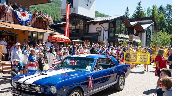A blue vintage car drives through a crowd of spectators at a sunny outdoor event, with onlookers, banners, and wooden buildings in the background.