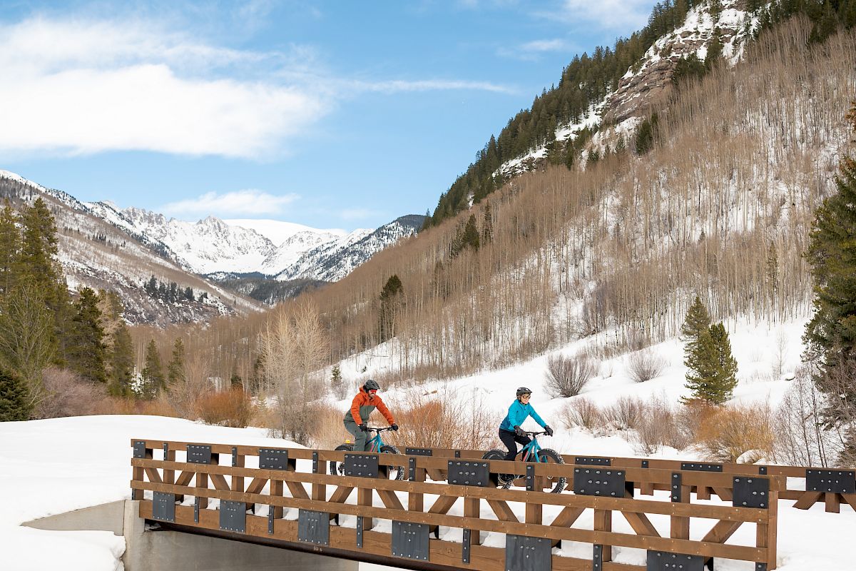 Two cyclists are riding across a wooden bridge in a snowy mountainous landscape with clear blue skies and scattered clouds in the background.