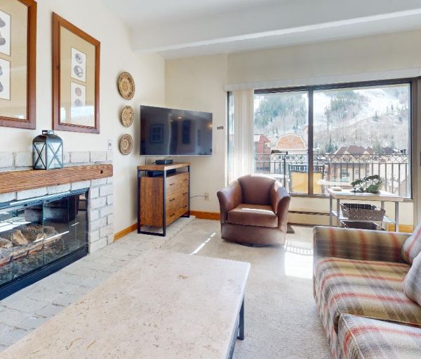 A cozy living room with a fireplace, plaid sofa, armchair, wall-mounted TV, artwork above the mantel, and large windows offering a snowy mountain view.