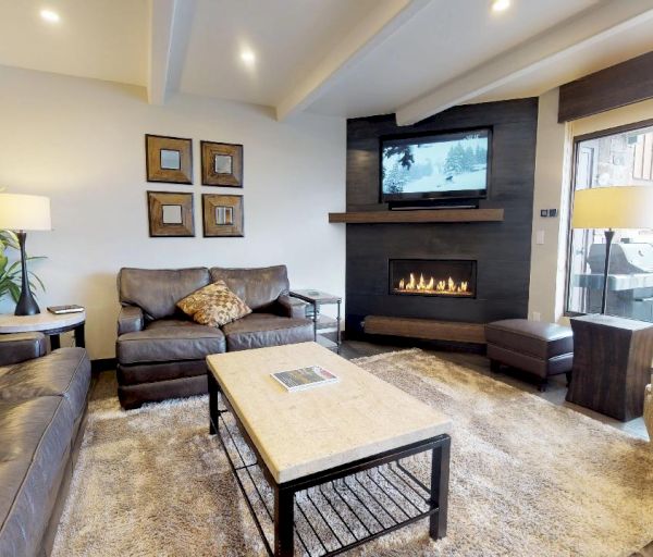 A cozy living room features modern furnishings, a fireplace, wall-mounted TV, and large window overlooking a balcony with outdoor view.
