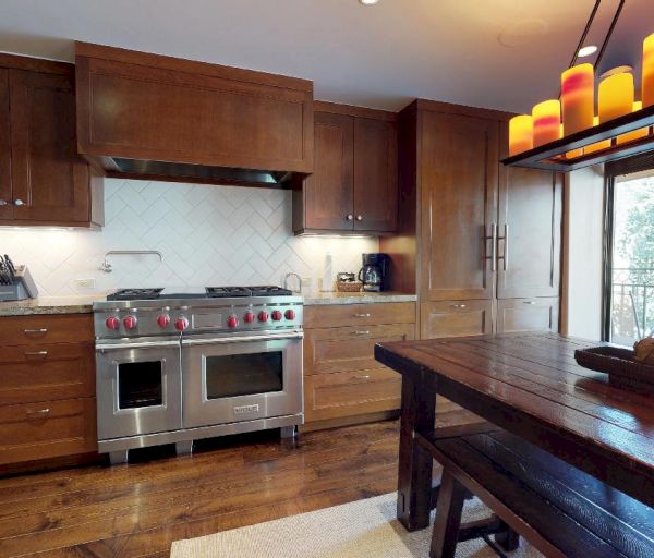 A modern kitchen with wooden cabinets, stainless steel appliances, a countertop, a wooden dining table, and large windows with a view outside.