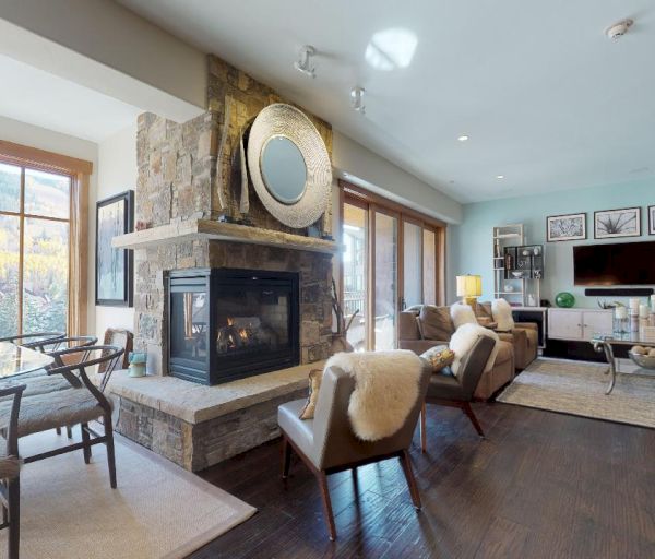 This image shows a modern living room with a stone fireplace, dining table, and large windows overlooking outdoor scenery.