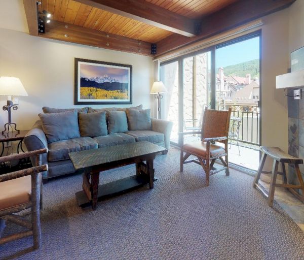 The image shows a cozy living room with a sofa, chairs, coffee table, fireplace, TV, lamps, and large windows looking out to a scenic view.