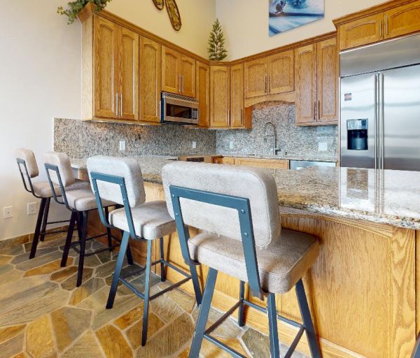 The image shows a kitchen with wooden cabinets, granite countertops, four upholstered barstools, a stainless steel refrigerator, and a stone tile floor.
