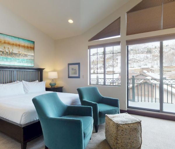 A well-lit bedroom with a large bed, two teal chairs, a small ottoman, dresser, bedside tables, artwork, and a window with a snowy view ending the sentence.