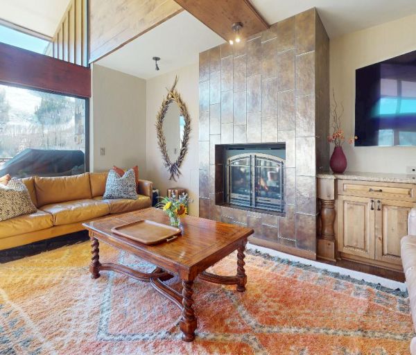 A cozy living room with large windows, two tan sofas, a fireplace, an area rug, a coffee table, a large TV, and wooden furniture, facing outdoors.
