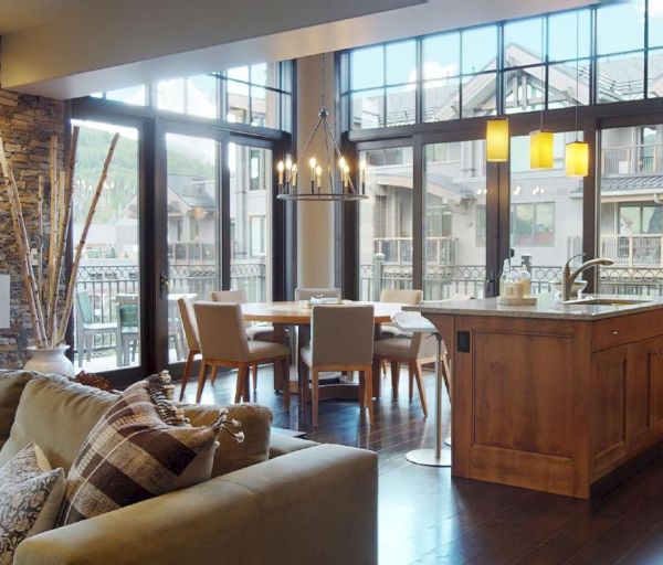 A cozy open-concept kitchen and living area with large windows, modern lighting, a stone fireplace, and wooden cabinetry and furniture.