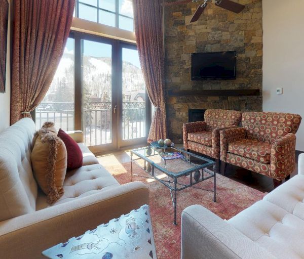 A cozy living room with two sofas, two patterned chairs, a coffee table, a wall-mounted TV, and a stone accent wall, with large windows and curtains.