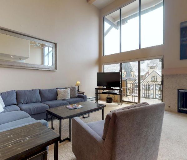 A cozy living room with a grey sectional sofa, a single armchair, a fireplace, a wall-mounted mirror, a TV, and large windows offering an outdoor view.