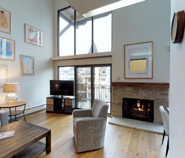 A cozy living room with high ceilings, large windows, a fireplace, a TV, artwork, and comfortable seating. There's a wooden coffee table, sofa, and chair.
