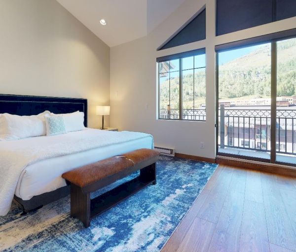 A modern bedroom features a large bed, nightstands with lamps, a bench, a blue rug, a dresser, and large windows with a mountain view outside.