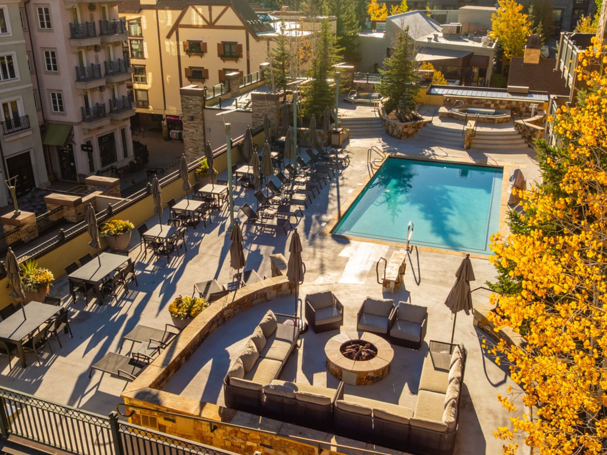 A sunny resort-style pool area with lounge chairs, umbrellas, a seating patio, and a rectangular pool surrounded by buildings and trees.
