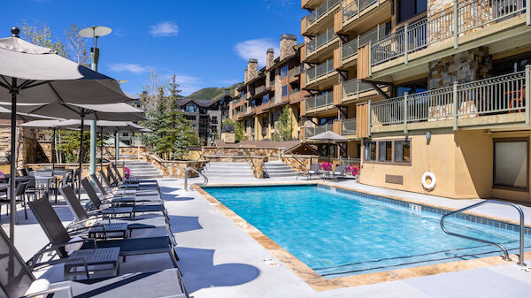 A sunny pool area at a resort with blue water, lounge chairs, umbrellas, and a multi-story beige building with balconies surrounding the pool.