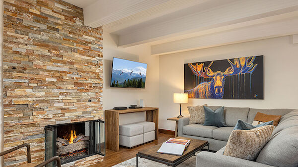 A cozy living room with a stone fireplace, a flat-screen TV, a gray sofa set, coffee table, and warm wall art in a bright, modern space.