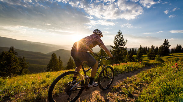 A person is riding a bicycle on a trail surrounded by greenery and trees, with the sun setting in the background, casting a warm glow.