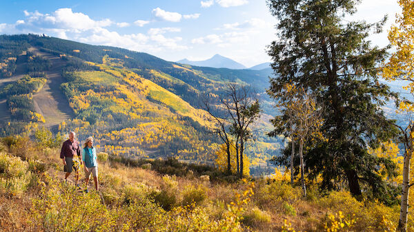 A couple strolls through a vibrant autumn landscape filled with colorful trees and mountains under a bright blue sky.